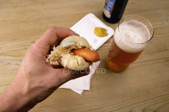 Cultivo mano masculina sosteniendo hot dog por encima del vaso de cerveza en la mesa - foto de stock