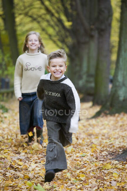 Boy running in front of girl along pathway covered by autumn leaves — Stock Photo