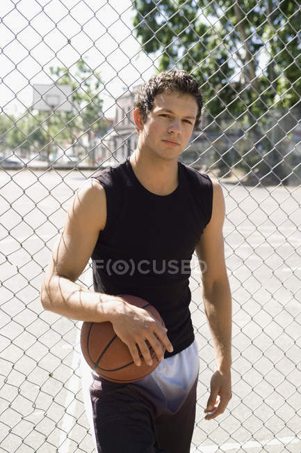 Un giovane uomo appoggiato ad una recinzione a catena che buca un pallone da basket — Foto stock