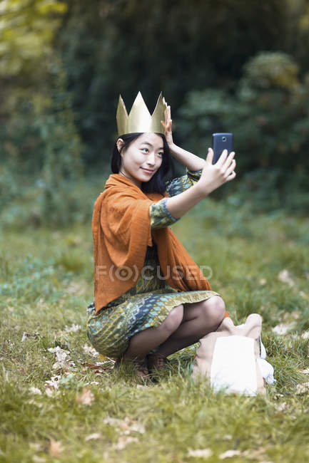 Портрет молодой женщины, одетой в корону и делающей селфи со смартфоном, сидящей на поле в парке — стоковое фото