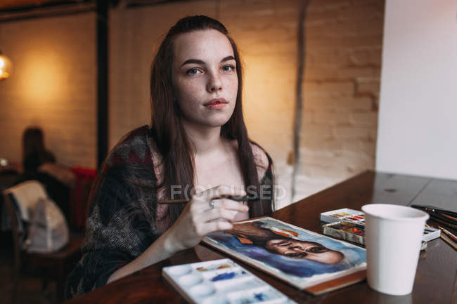 Portrait of young woman sitting by painting on table at cafe — Stock Photo