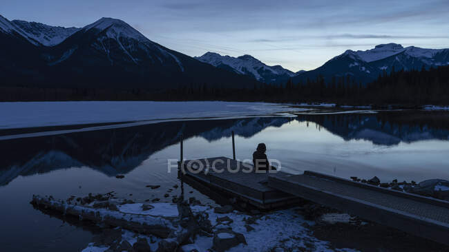 Homem sentado no cais tranquilo lago de inverno com vista montanha à noite, Banff, Alberta, Canadá — Fotografia de Stock
