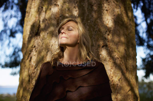 Portrait of woman with eyes closed standing at sunny tree trunk — Stock Photo