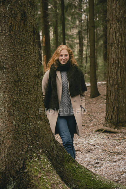 Portrait confident redhead woman in coat and scarf standing among trees in woods — Stock Photo