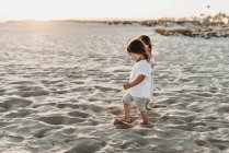 Вид сбоку маленьких девочек, держащихся за руки и гуляющих на пляже — стоковое фото