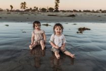 Vista frontal de hermanas pequeñas sentadas en el agua en la playa - foto de stock