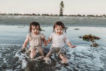 Vista frontal de irmãs crianças sentadas e espirrando na água na praia — Fotografia de Stock