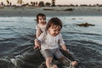 Frontansicht von Kleinkind-Schwestern, die am Strand im Wasser sitzen und planschen — Stockfoto