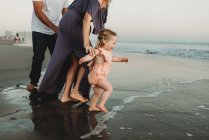 Vue latérale de la jeune fille avec la famille courant dans l'océan au coucher du soleil — Photo de stock