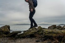 Uomo con zaino sale piccolo sentiero di rocce accanto baia sotto cielo grigio — Foto stock