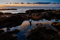 Adolescent marche sur rocheux rivage coulée réflexion sous coucher de soleil ciel — Photo de stock