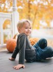 A little boy sitting on a porch by orange maple trees and pumpkins — Stock Photo