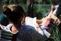Mulher com óculos de sol lendo um livro deitado em uma rede. — Fotografia de Stock