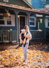 Una mujer con un niño está de pie cerca de una casa en hojas amarillas - foto de stock