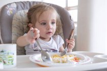 Bambina che mangia da sola sulla sedia vicino al tavolo. — Foto stock