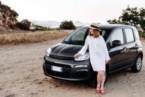 Mignon, charmante fille dans un chapeau de paille pose autour d'une voiture noire sur le bord de la route. — Photo de stock