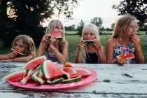 Братья и сёстры сидят за столом для пикника и едят арбуз летом — стоковое фото