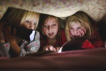 Três meninas lendo livro sob cobertor com lanterna — Fotografia de Stock