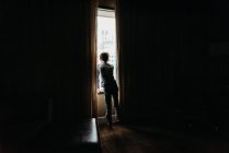 Young boy looking out a window of a darkened room at tall buildings. — Stock Photo