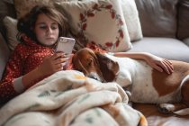 Schönes Mädchen mit Smartphone entspannt sich im Bett mit ihrem Hund — Stockfoto