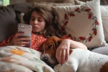 Красива дівчина з смартфоном розслабляється в ліжку зі своєю собакою — стокове фото