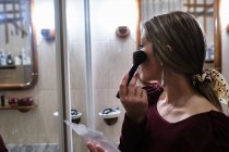 Mujer joven se pone maquillaje en el baño mientras se mira en el espejo - foto de stock