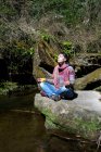 Young woman practicing meditation on a riverside stone. — Stock Photo