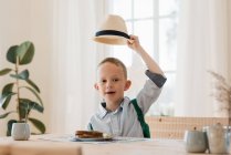 Boy holding his hat up whilst eating his lunch at home smiling — Stock Photo