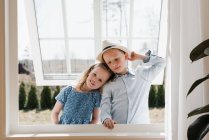 Brother and sister hugging whilst looking through a window at home — Stock Photo