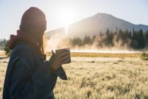 Молода жінка тримає каву на сході сонця, дивлячись на туманну гору — стокове фото