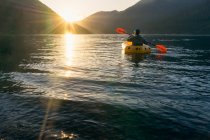 Jovem remando no lago ao pôr do sol em direção às montanhas — Fotografia de Stock