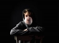 Giovanissima ragazzo in hoodie seduta su un sedia in buio stanza soffiando un bolla. — Foto stock