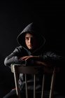 Portrait d'un adolescent en sweat à capuche assis sur une chaise dans une pièce sombre. — Photo de stock