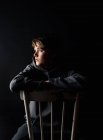 Портрет мальчика-подростка, сидящего на стуле в темной комнате. — стоковое фото
