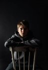 Низький ключовий портрет хлопчика-підлітка, що сидить на стільці в темній кімнаті . — стокове фото