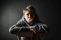 Adolescente menino com capuz sentado em uma cadeira no quarto escuro soprando uma bolha. — Fotografia de Stock