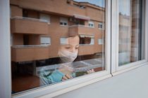 Маленькая девочка в маске смотрит в окно, запертое дома пандемией коронавируса ковида-19. — стоковое фото