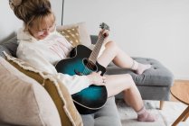 Woman sat playing guitar at home on the couch — Stock Photo