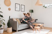 Daughter kissing her mom whilst she plays guitar at home — Stock Photo