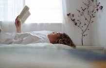 Mulher de cabelos encaracolados lendo um livro deitado na cama — Fotografia de Stock