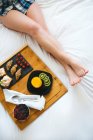 Primer plano de piernas femeninas y bandeja de desayuno en la cama en el dormitorio blanco - foto de stock
