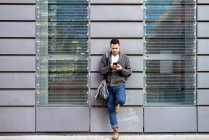 Front view of a bearded man using phone leaning on office building wall — Stock Photo
