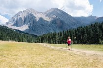 Wanderin mit Strohhut, kurzer Hose und Rucksack auf dem Weg über eine Ebene mit herrlichen Bergen im Hintergrund, während sie die Natur ringsum genießt. Horizontales Foto. — Stockfoto