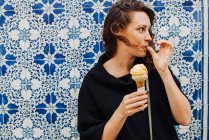 Milenial mujer lamiendo dedo mientras come helado en una pared de azulejos - foto de stock