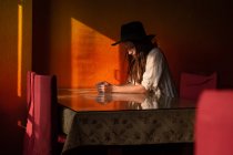 Retrato de una joven mujer caucásica reflexiva en sombrero negro sentado junto a la mesa en la cafetería solo frente a la pared naranja - foto de stock