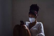 Молодая женщина в маске с помощью смартфона в темной комнате — стоковое фото