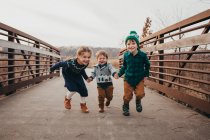 Drei Geschwister halten sich Händchen und rennen auf Brücke in Richtung Kamera — Stockfoto