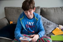 Souriant adolescent garçon lit carte d'anniversaire tout en étant assis sur canapé à Pjs — Photo de stock