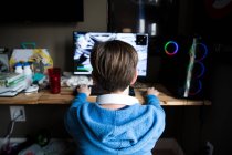 Vista trasera de adolescente chico jugando juego de ordenador en un escritorio desordenado - foto de stock