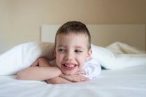 Adorable smiling kid lying down on bed under blanket, looking camera — Stock Photo